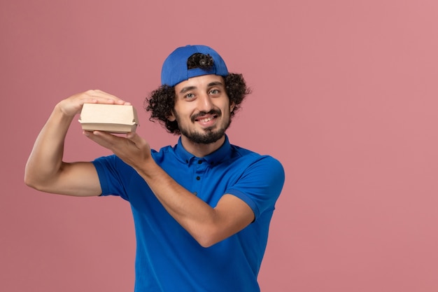 Mensajero masculino de vista frontal en uniforme azul y capa sosteniendo un pequeño paquete de comida de entrega en la pared rosa