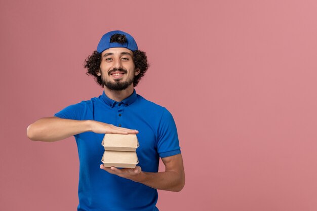 Mensajero masculino de vista frontal en uniforme azul y capa sosteniendo pequeños paquetes de comida de entrega en la pared rosa