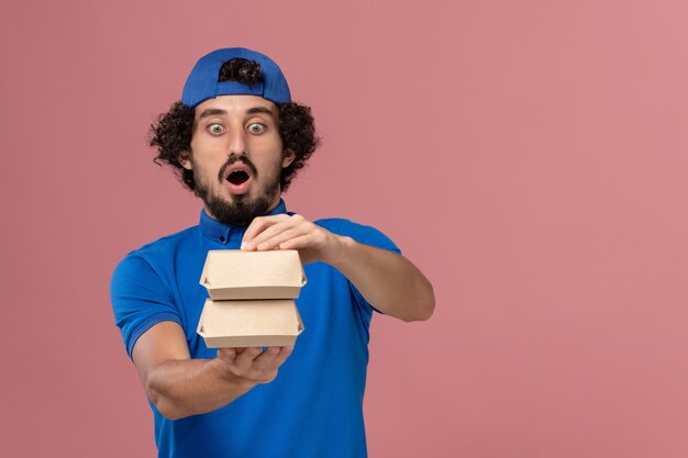 Mensajero masculino de vista frontal en uniforme azul y capa sosteniendo pequeños paquetes de comida de entrega en la pared rosa