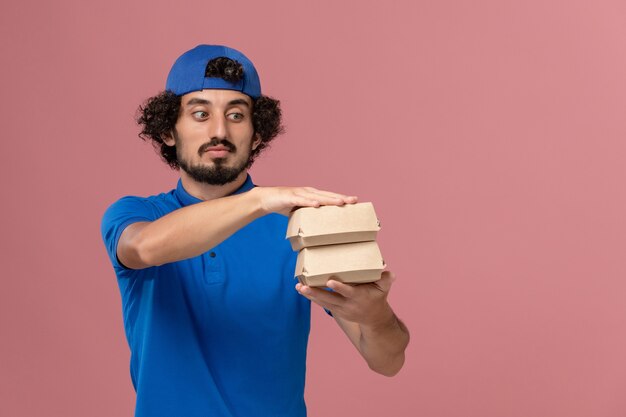 Mensajero masculino de vista frontal en uniforme azul y capa sosteniendo pequeños paquetes de comida de entrega en la pared rosa