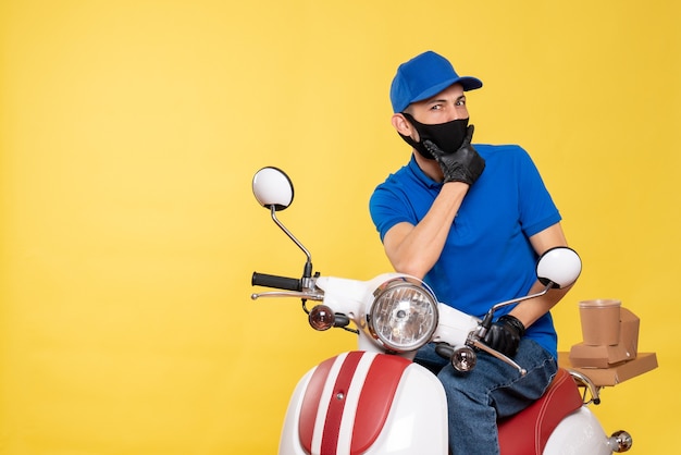 Mensajero masculino de vista frontal en uniforme azul y máscara en la bicicleta amarilla covid- servicio de virus de trabajo de entrega de trabajo pandémico