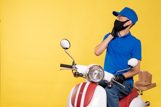 Mensajero masculino de vista frontal en uniforme azul y máscara en la bicicleta amarilla covid- servicio de virus de trabajo de entrega de trabajo pandémico