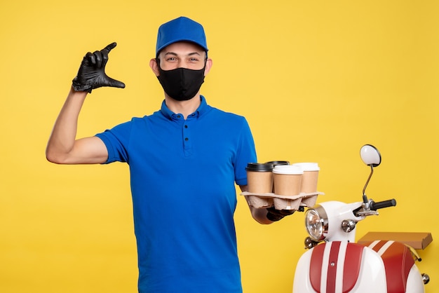 Mensajero masculino de vista frontal en uniforme azul y máscara sosteniendo café en un uniforme de trabajo amarillo covid- servicio de trabajo pandemia de entrega