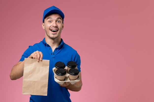 Mensajero masculino de vista frontal en uniforme azul con paquete de alimentos y café en la pared rosa, entrega de trabajo de servicio uniforme