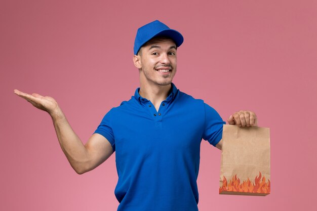 Mensajero masculino de vista frontal en uniforme azul con paquete de alimentos de papel sonriendo en la pared rosa, entrega de servicio uniforme de trabajador de trabajo