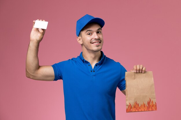 Mensajero masculino de vista frontal en uniforme azul con paquete de alimentos de papel con tarjeta en la pared rosa, entrega de servicio uniforme de trabajador