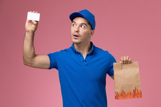 Mensajero masculino de vista frontal en uniforme azul con paquete de alimentos con tarjeta en la pared rosa, entrega de servicio uniforme de trabajo