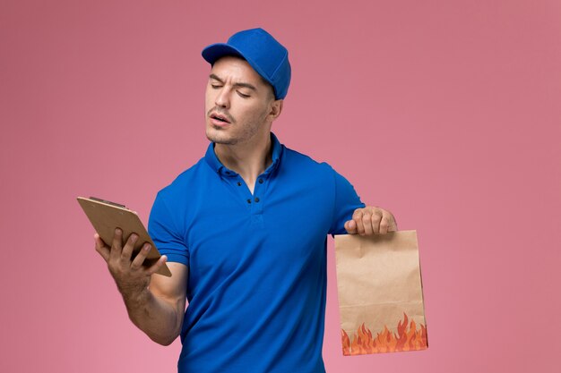 Mensajero masculino de vista frontal en uniforme azul con paquete de comida de bloc de notas en la pared rosa, entrega de trabajo de servicio uniforme