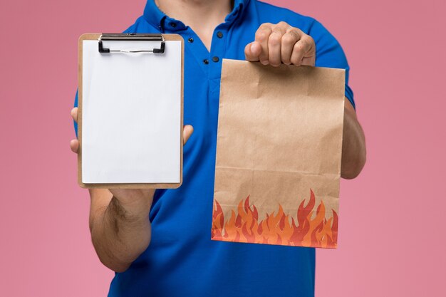 Mensajero masculino de vista frontal en uniforme azul con paquete de comida de papel y bloc de notas en la pared rosa, entrega de trabajo de servicio uniforme