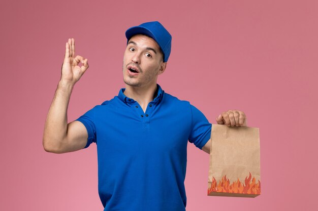 Mensajero masculino de vista frontal en uniforme azul con paquete de comida de papel en la pared rosa, entrega de trabajo de servicio uniforme de trabajador