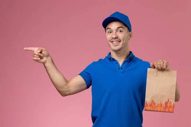 Mensajero masculino de vista frontal en uniforme azul con paquete de papel con sonrisa en la pared rosa, entrega de servicio uniforme de trabajador de trabajo