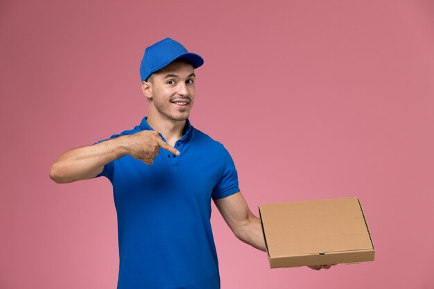 Mensajero masculino de vista frontal en uniforme azul sosteniendo la apertura de la caja de entrega de alimentos en la pared rosa, entrega de servicio de trabajo uniforme