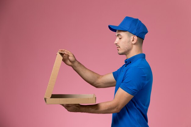 Mensajero masculino de vista frontal en uniforme azul sosteniendo la apertura de la caja de entrega de alimentos en la pared rosa, entrega de trabajo de servicio uniforme