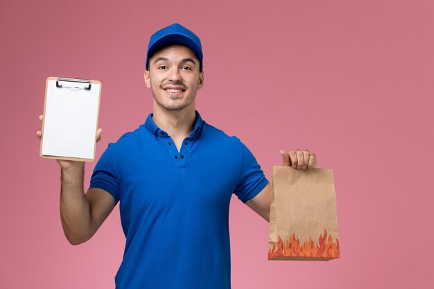 Mensajero masculino de vista frontal en uniforme azul sosteniendo el paquete de alimentos con el bloc de notas en la pared rosa, entrega de servicio uniforme de trabajador de trabajo