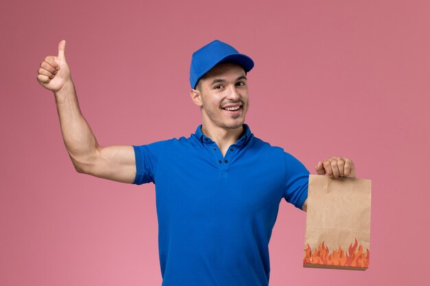 Mensajero masculino de vista frontal en uniforme azul sosteniendo el paquete de alimentos que muestra como un letrero en la pared rosa, entrega de servicio uniforme de trabajador de trabajo