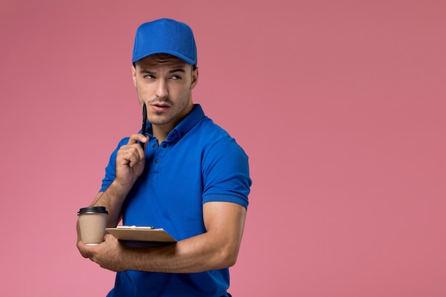 Mensajero masculino de vista frontal en uniforme azul sosteniendo un pequeño bloc de notas y café pensando en la pared rosa, entrega de servicio uniforme del trabajador