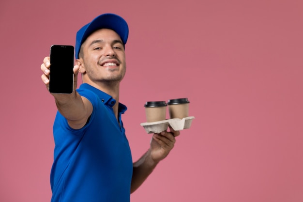 Foto gratuita mensajero masculino de vista frontal en uniforme azul sosteniendo su teléfono y tazas de café en la pared rosa, entrega de trabajo de servicio uniforme