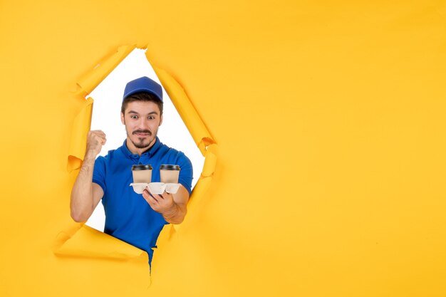Mensajero masculino de vista frontal en uniforme azul sosteniendo tazas de café en el espacio amarillo