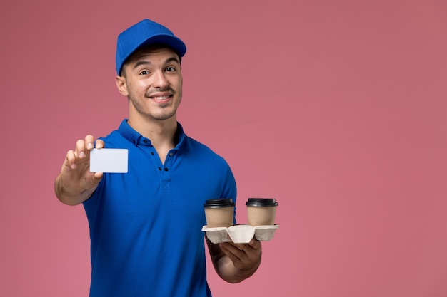 Mensajero masculino de vista frontal en uniforme azul sosteniendo tazas de café de tarjeta blanca con sonrisa en la pared rosa, entrega de servicio uniforme de trabajador de trabajo