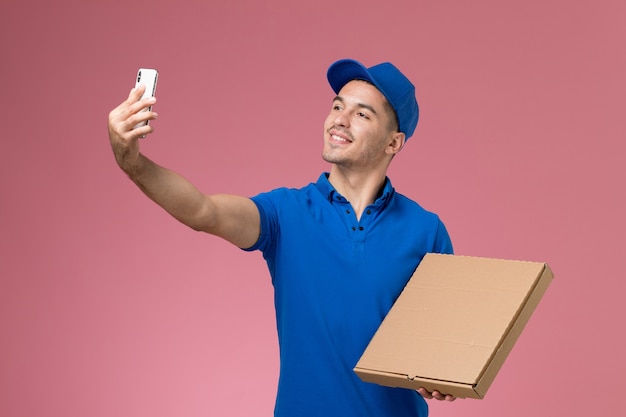 Mensajero masculino de vista frontal en uniforme azul tomando selfie con caja de comida en la pared rosa, entrega de trabajo de servicio uniforme