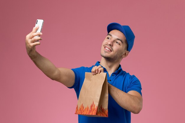 Mensajero masculino de vista frontal en uniforme azul tomando un selfie con paquete de comida en la pared rosa, entrega de servicio de trabajador uniforme