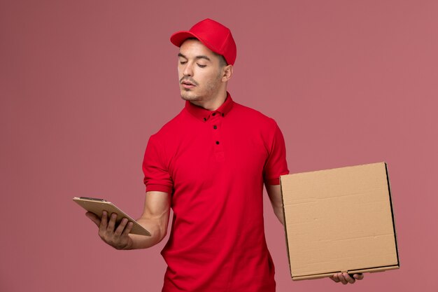 Mensajero masculino de vista frontal en uniforme rojo y capa con caja de comida de bloc de notas en la pared rosa trabajo de servicio uniforme de reparto masculino