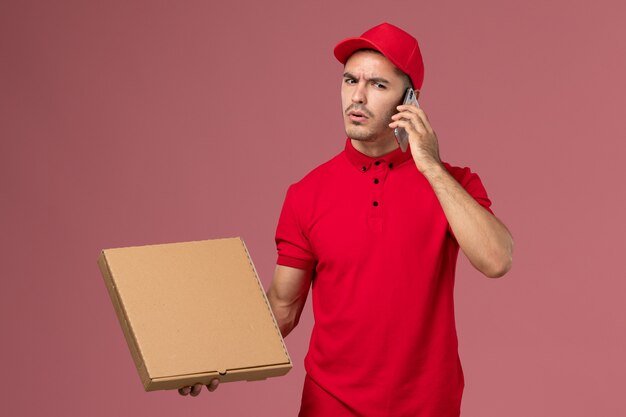 Mensajero masculino de vista frontal en uniforme rojo y capa con caja de comida hablando por teléfono en el escritorio rosa
