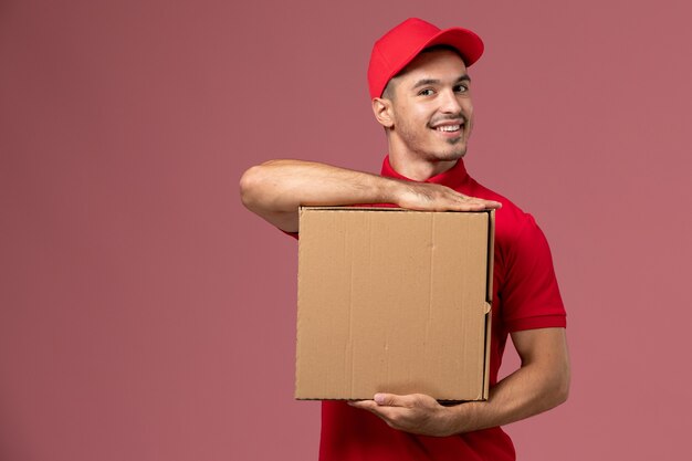 Mensajero masculino de vista frontal en uniforme rojo y capa con caja de comida con una sonrisa en el trabajo de trabajador de pared rosa