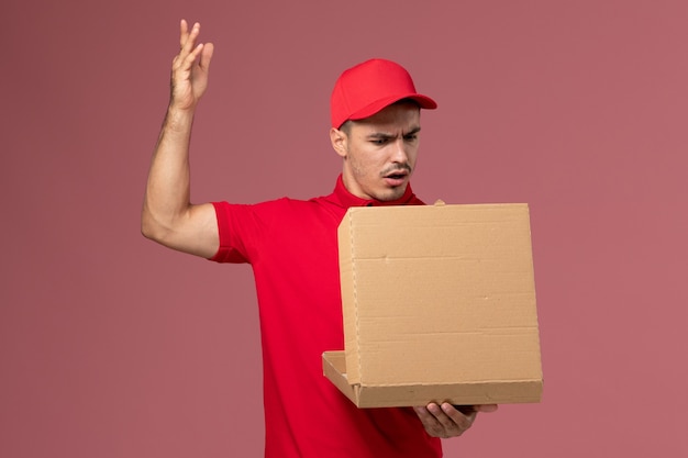 Mensajero masculino de vista frontal en uniforme rojo y capa con caja de comida en el trabajador de la pared de color rosa claro