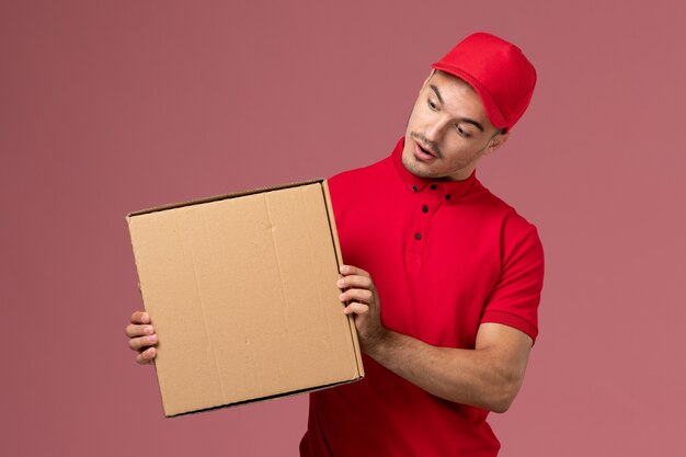 Mensajero masculino de vista frontal en uniforme rojo y capa con caja de comida en el trabajo de trabajador de pared rosa