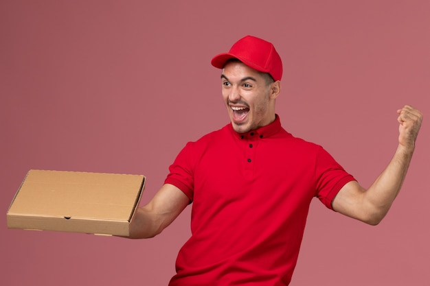 Mensajero masculino de vista frontal en uniforme rojo y capa con caja de entrega de alimentos regocijándose en el trabajo de trabajador de pared rosa