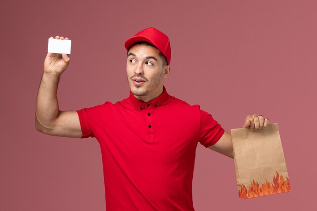 Mensajero masculino de vista frontal en uniforme rojo y capa con paquete de alimentos y tarjeta en el uniforme de trabajo de trabajador de entrega de servicio de escritorio rosa