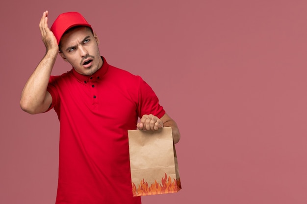 Mensajero masculino de vista frontal en uniforme rojo y capa con paquete de comida de papel pensando en el trabajador de la pared rosa