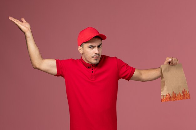 Mensajero masculino de vista frontal en uniforme rojo y capa con paquete de comida de papel en el trabajador de la pared rosa