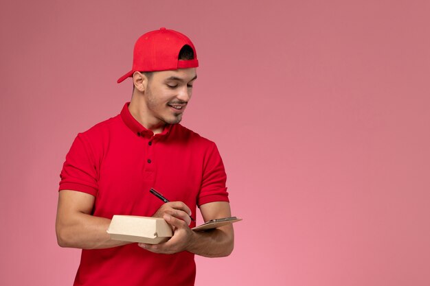 Mensajero masculino de la vista frontal en uniforme rojo y capa que sostiene el pequeño paquete de entrega con el bloc de notas escribiendo una nota sobre fondo rosa.