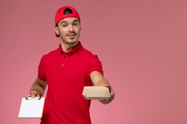 Mensajero masculino de la vista frontal en uniforme rojo y capa que sostiene el pequeño paquete de entrega con el bloc de notas escribiendo una nota sobre fondo rosa.