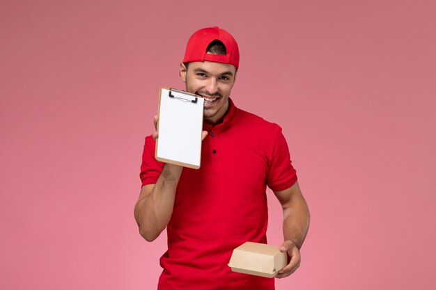 Mensajero masculino de la vista frontal en uniforme rojo y capa que sostiene el pequeño paquete de entrega con el bloc de notas en el fondo rosa.