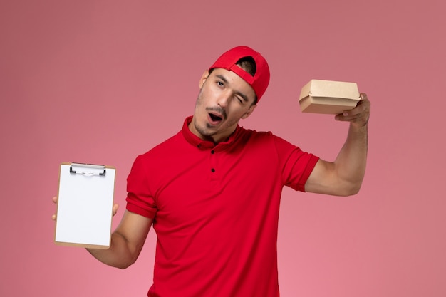 Mensajero masculino de la vista frontal en uniforme rojo y capa que sostiene el pequeño paquete de entrega con el bloc de notas en el fondo rosa.