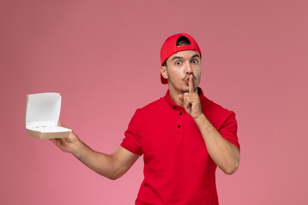 Mensajero masculino de la vista frontal en uniforme rojo y capa que sostiene el pequeño paquete de entrega en el escritorio rosa.