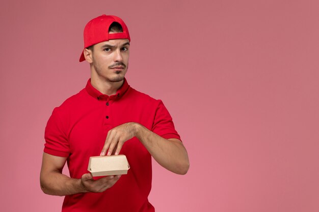 Mensajero masculino de la vista frontal en uniforme rojo y capa que sostiene el pequeño paquete de entrega en el fondo rosa.