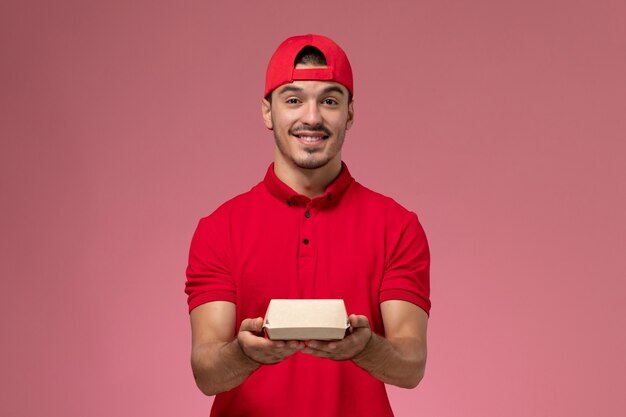 Mensajero masculino de la vista frontal en uniforme rojo y capa que sostiene el pequeño paquete de entrega en el fondo rosa.