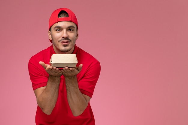 Foto gratuita mensajero masculino de la vista frontal en uniforme rojo y capa que sostiene el pequeño paquete de entrega en el fondo rosa.