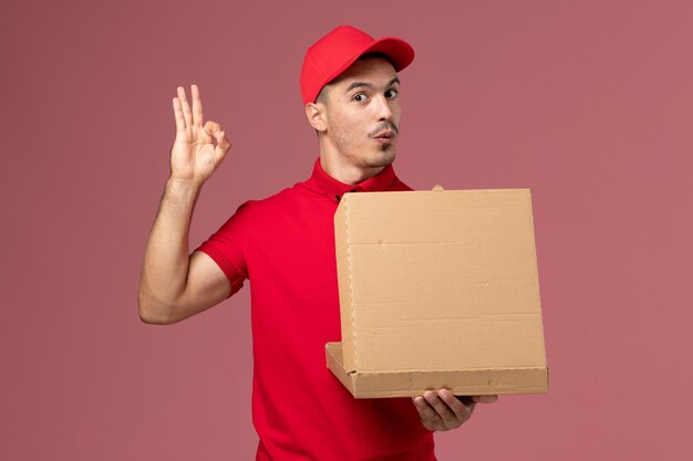 Mensajero masculino de vista frontal en uniforme rojo y capa sosteniendo la caja de comida y abriéndola en la pared de color rosa claro