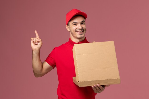 Mensajero masculino de vista frontal en uniforme rojo y capa sosteniendo la caja de comida y abriéndola sonriendo en el trabajo de trabajador de pared rosa claro