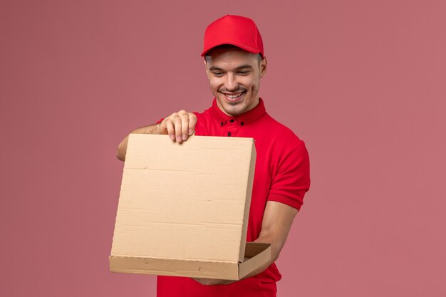 Mensajero masculino de vista frontal en uniforme rojo y capa sosteniendo la caja de comida y abriéndola en el trabajo de trabajador de pared rosa claro