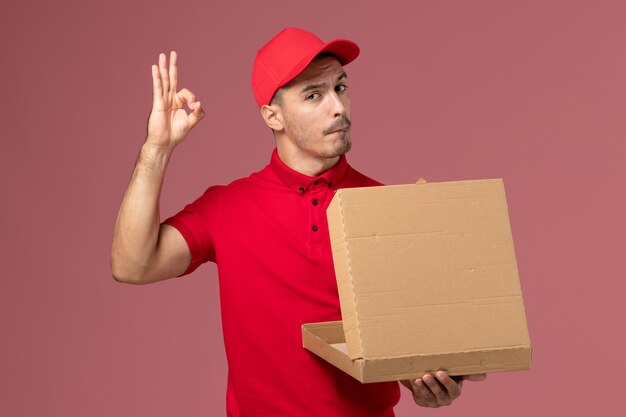 Mensajero masculino de vista frontal en uniforme rojo y capa sosteniendo la caja de comida y abriéndola en el trabajo de trabajador de pared rosa claro