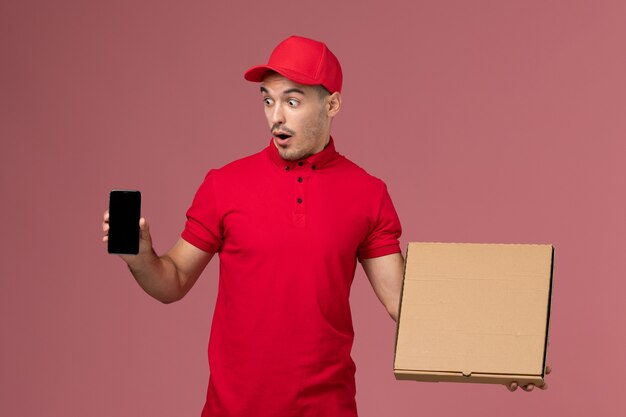 Mensajero masculino de vista frontal en uniforme rojo y capa sosteniendo la caja de comida junto con el teléfono en el uniforme de entrega masculino de trabajo de servicio de pared rosa