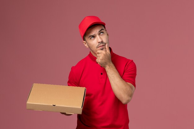 Mensajero masculino de vista frontal en uniforme rojo y capa sosteniendo la caja de entrega de alimentos pensando en el varón trabajador de escritorio rosa