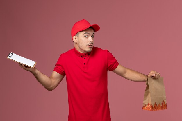 Mensajero masculino de vista frontal en uniforme rojo y capa sosteniendo el paquete de alimentos y el bloc de notas en la pared rosa