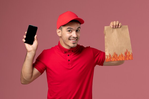 Mensajero masculino de vista frontal en uniforme rojo y capa sosteniendo el paquete de alimentos y el teléfono en la pared rosa